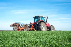 Red Chinese farm tractor pulling a plow in a green agricultural field.